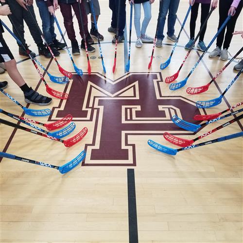 Students standing around a circle, holding their floor ball sticks on the floor 